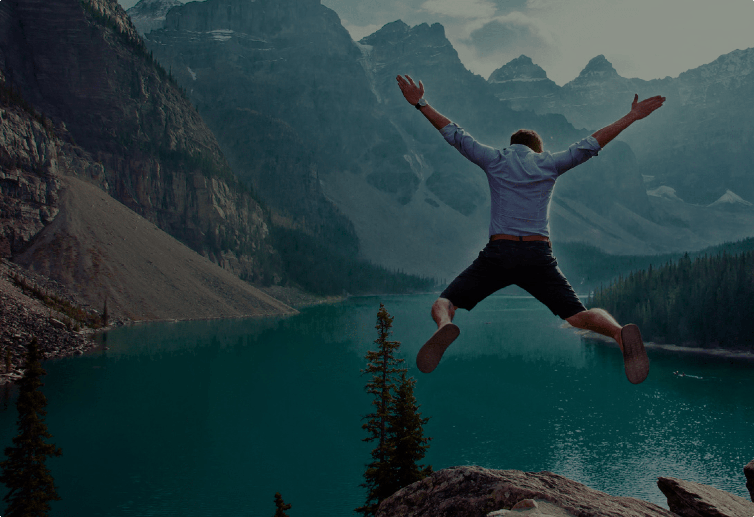 Person jumping with arms raised on a rocky cliff overlooking a turquoise mountain lake surrounded by tall pine trees and dramatic, rugged peaks.
